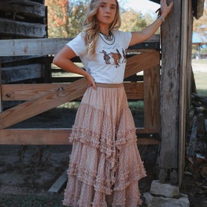 May include: A woman wearing a white t-shirt with a cowboy graphic, a tiered blush pink skirt, and cow-print boots. The skirt has a dotted pattern and a ruffled design. The woman is standing near a wooden fence.