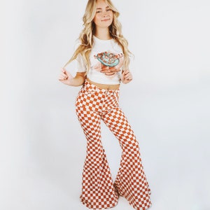 Può includere: Una donna indossa un crop top bianco con una grafica e pantaloni a zampa a scacchi arancioni e bianchi. La parte superiore ha una grafica di un personaggio. I pantaloni hanno una vita alta e un'ampia svasatura.