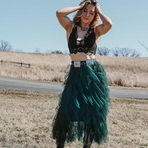 Pode incluir: Um top de renda preta combinado com uma saia de tule verde escuro e um cinto prateado. O traje é complementado com botas de cowboy pretas. O fundo apresenta um campo e um céu azul claro, criando um visual boêmio elegante.