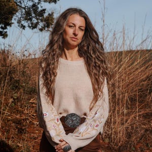 May include: A beige sweater with floral embroidery on the sleeves. The sweater is paired with brown pants and a silver belt buckle. The person is also wearing a silver ring. The background features tall, dry grass and a clear sky.