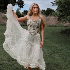 May include: A sleeveless corset top featuring a multi-colored floral pattern on a cream background, paired with a long, sheer, off-white skirt. The skirt has a tiered design with ruffled edges and a lightweight, flowing fabric construction.