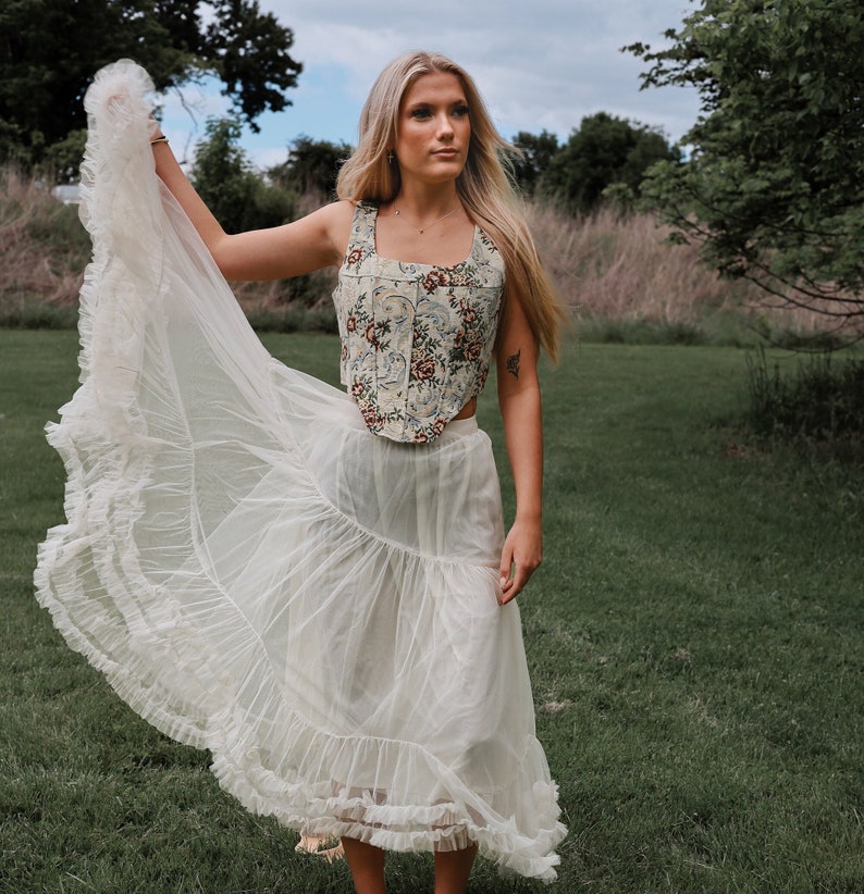 May include: A sleeveless corset top featuring a multi-colored floral pattern on a cream background, paired with a long, sheer, off-white skirt. The skirt has a tiered design with ruffled edges and a lightweight, flowing fabric construction.