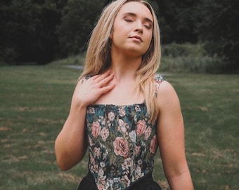 May include: A woman wearing a black and green floral print corset top with a black skirt. The corset top has a square neckline and is sleeveless.