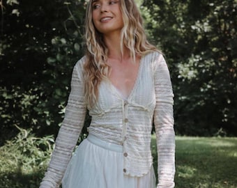 May include: A long-sleeve cream-colored lace cardigan featuring a V-neckline and a front button closure, paired with a layered white tulle skirt. The cardigan has a textured, semi-sheer pattern throughout the fabric and fitted sleeves.
