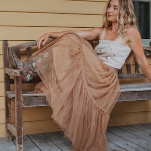 Könnte beinhalten: Outfit einer Frau mit einem beigefarbenen Crop-Top mit floraler Stickerei und einem langen, fließenden, hellbraunen Tüllrock. Dazu braune Cowboy-Stiefel mit floraler Stickerei. Die Frau sitzt auf einer Holzbank.