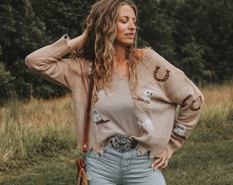 May include: A woman wearing a light brown sweater with a white horseshoe and cowboy hat design. The sweater has a frayed hem and is worn with a brown leather belt and blue jeans.