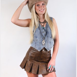 May include: A brown faux leather skirt with pleats and pockets. The skirt is paired with a light blue denim vest and a layered silver necklace with turquoise accents.