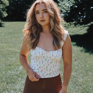 May include: A woman wearing a white floral print sleeveless top with a brown pair of pants.