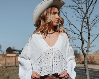 May include: A white crochet top with a long sleeve and a button closure. The top has a lace trim and is worn with a brown leather skirt and a white cowboy hat.