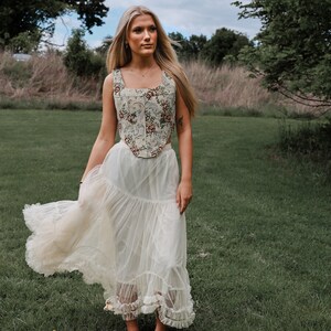 May include: A sleeveless corset top featuring a cream-colored base with a multi-colored floral and bird pattern, paired with a long, flowing, off-white tulle skirt that has a tiered design and ruffled hem detailing at the bottom of the garment.