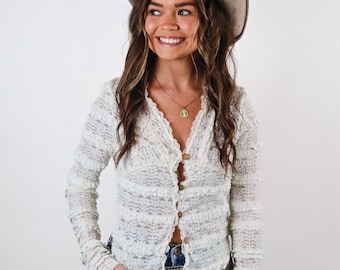 May include: A cream-colored, long-sleeved cardigan with a textured, open-knit design. The button-down shirt has a V-neck and is paired with blue jeans and a brown cowboy hat. A gold necklace with a pendant is also visible.