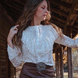 May include: A long-sleeved white lace blouse featuring a button-down front, a round neckline, and layered ruffled detailing across the chest. The blouse is paired with a dark brown belt that has a large, ornate, circular metal buckle.