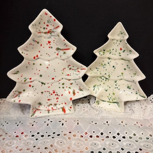 Puede incluir: Dos platos de cerámica con forma de árbol de Navidad. Uno es blanco con motas rojas y verdes, el otro es blanco con motas verdes. Los platos están sobre una superficie blanca con ojales sobre un fondo negro. Decoración festiva navideña.