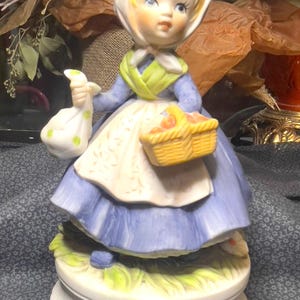 Può includere: Una statuetta in porcellana di una ragazza con un vestito blu e un grembiule bianco, che tiene un cesto di frutta e una borsa. Indossa un berretto bianco e ha i capelli biondi. La statuetta è su una base bianca con un disegno di erba verde.