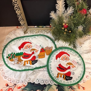 May include: Christmas-themed placemat and coaster set featuring Santa Claus holding a menu. The placemat is oval, and the coaster is round, both with a white background, green trim, and a scalloped edge. The text "Holiday Menu" is visible.