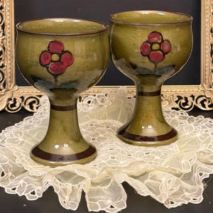 May include: Two olive green ceramic goblets with a red floral design. The goblets have a rounded bowl shape and a stem with a dark brown band. The goblets are displayed on a white lace doily.