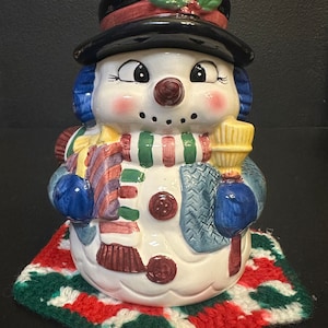 May include: A ceramic snowman figurine with a black top hat, blue scarf, and a red and green knitted hat. The snowman is holding a yellow broom and is sitting on a red, green, and white knitted mat.