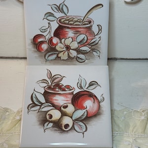 Pode incluir: Dois azulejos de cerâmica vintage com imagens pintadas à mão de frutas e flores. O azulejo superior apresenta uma panela vermelha com uma colher, cerejas e uma flor branca. O azulejo inferior apresenta uma panela vermelha com bagas, uma maçã vermelha e bagas brancas.