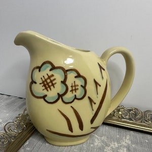 May include: A vintage cream-colored ceramic pitcher with a brown and blue floral design. The pitcher has a rounded body and a handle.