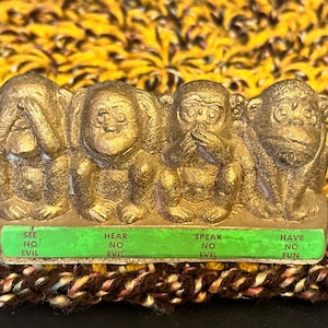 May include: A gold-colored figurine depicting the three wise monkeys: see no evil, hear no evil, and speak no evil. The monkeys are arranged side by side on a green base with text. The figurine is on a textured background.