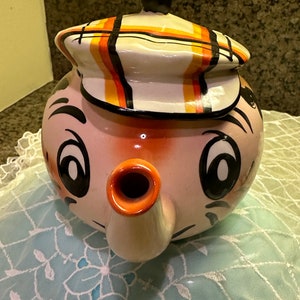May include: A ceramic teapot shaped like a cartoon face with a black and orange striped hat. The teapot has a large orange spout and black eyes.