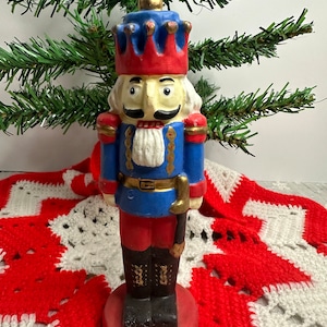 Puede incluir: Una vela vintage de cascanueces con forma de soldado. La vela está pintada de azul, rojo y dorado con barba y bigote blancos. La vela está sobre una base roja.