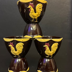 May include: A stack of three brown ceramic egg cups. Each cup features a yellow rooster design with red accents. The rims and bases of the cups are also yellow. The egg cups are stacked in a pyramid shape.