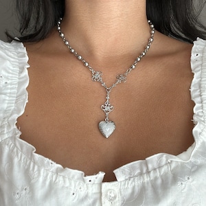 May include: Silver-toned necklace featuring a beaded chain and a decorative pendant. The pendant includes a heart-shaped locket with intricate detailing. The necklace is worn against a white eyelet top, creating a contrast.