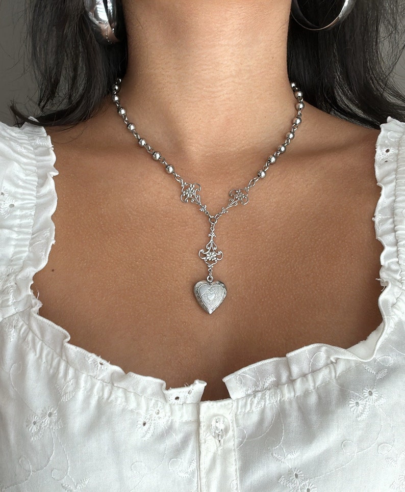 May include: Silver-toned necklace featuring a beaded chain and a decorative pendant. The pendant includes a heart-shaped locket with intricate detailing. The necklace is worn against a white eyelet top, creating a contrast.