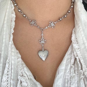 May include: Silver-toned necklace featuring a heart-shaped locket pendant. The necklace has a delicate design with ornate filigree accents and a beaded chain. The locket is detailed with engraved patterns, creating a vintage aesthetic.