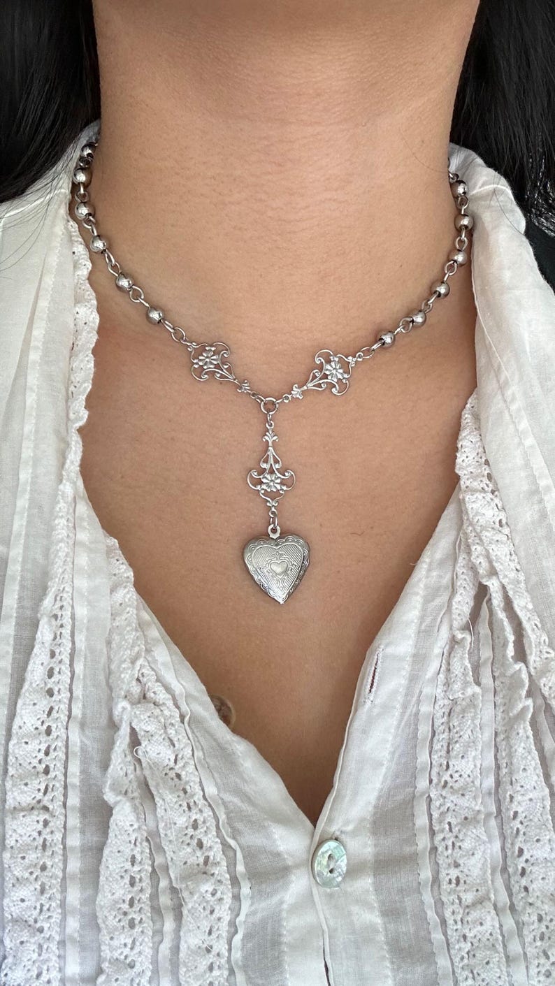 May include: Silver-toned necklace featuring a heart-shaped locket pendant. The necklace has a delicate design with ornate filigree accents and a beaded chain. The locket is detailed with engraved patterns, creating a vintage aesthetic.