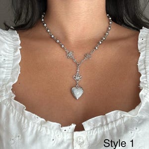 May include: A silver-toned necklace with a heart-shaped pendant. The necklace features a beaded chain and ornate floral details. The pendant is a locket with a textured surface. The text "Style 1" is visible.