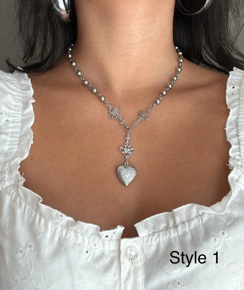 May include: A silver-toned necklace with a heart-shaped pendant. The necklace features a beaded chain and ornate floral details. The pendant is a locket with a textured surface. The text "Style 1" is visible.