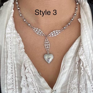 May include: A silver-toned necklace with a heart-shaped locket pendant. The necklace features a delicate chain with small beads and ornate filigree details. The text "Style 3" is displayed in black.