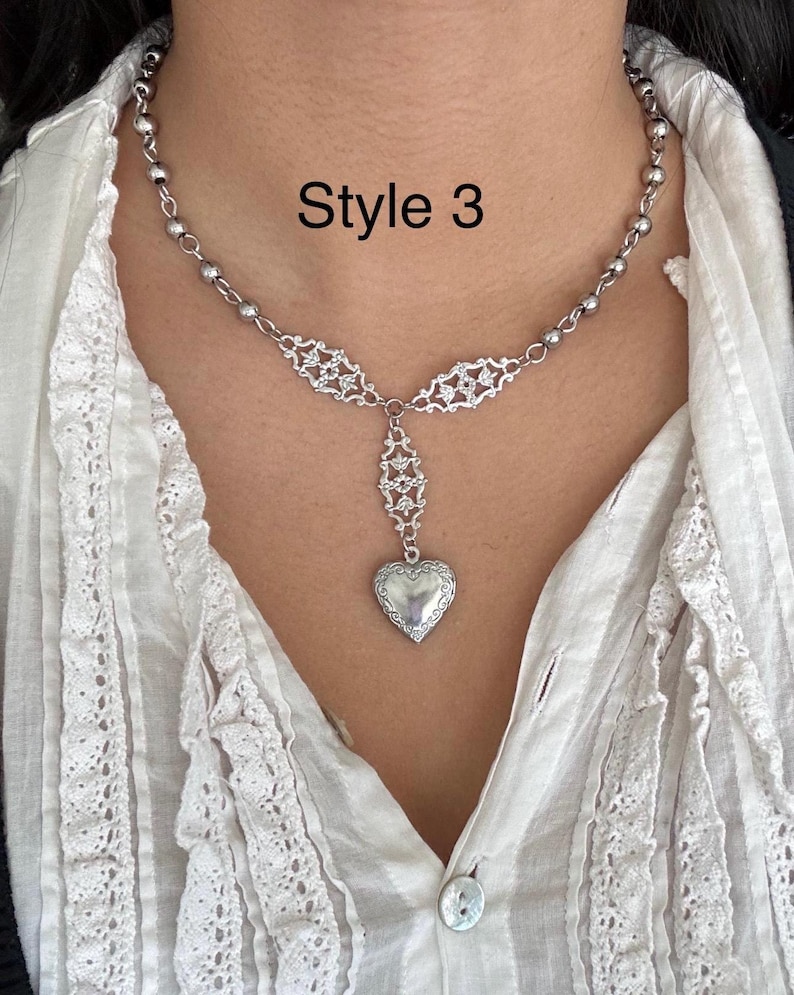 May include: A silver-toned necklace with a heart-shaped locket pendant. The necklace features a delicate chain with small beads and ornate filigree details. The text "Style 3" is displayed in black.