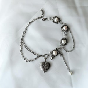Puede incluir: Un collar de cadena de plata con un colgante en forma de corazón y detalles de perlas. El collar tiene un diseño delicado y femenino.