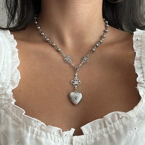 May include: Silver-toned necklace featuring a beaded chain and a decorative pendant. The pendant has a heart-shaped locket design with intricate detailing. The necklace is worn against a white blouse.