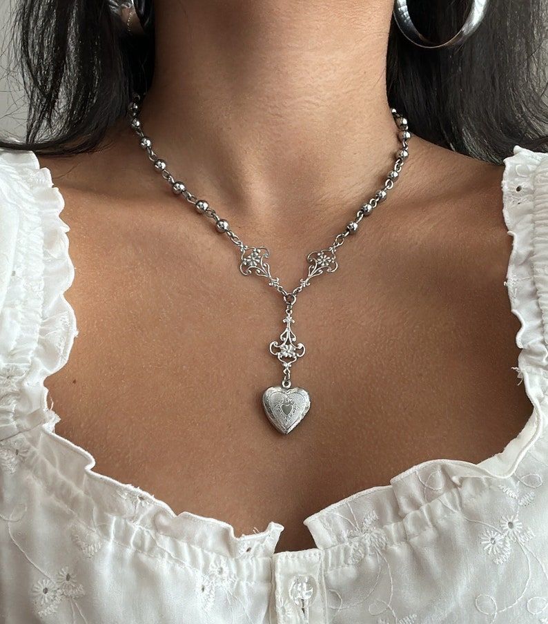 May include: Silver-toned necklace featuring a beaded chain and a decorative pendant. The pendant has a heart-shaped locket design with intricate detailing. The necklace is worn against a white blouse.