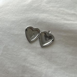 May include: An open, silver-colored heart-shaped locket. The locket is on a white surface. The inside of the locket has a decorative design. The locket is open, revealing the inside of the heart.