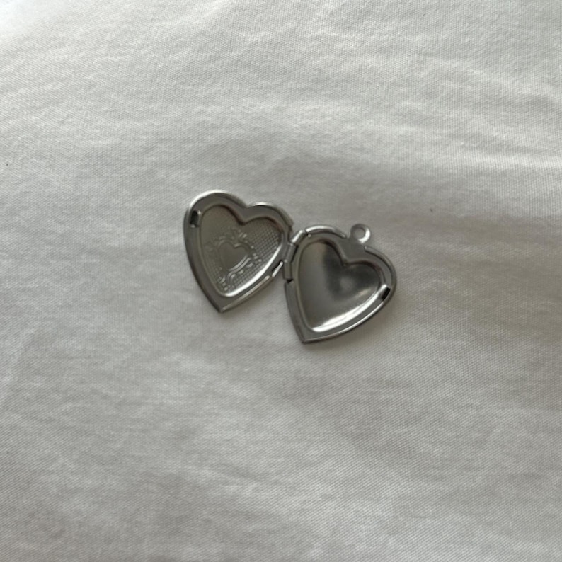 May include: An open, silver-colored heart-shaped locket. The locket is on a white surface. The inside of the locket has a decorative design. The locket is open, revealing the inside of the heart.
