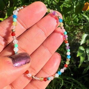 May include: A delicate beaded bracelet featuring small, multi-coloured faceted gemstone beads in shades of red, blue, yellow, green, purple, and clear, interspersed with tiny, polished gold-tone metallic spacer beads arranged in a circular pattern.