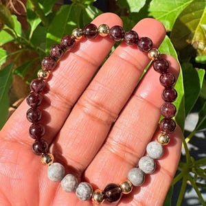 May include: A bracelet with a mix of dark red, grey, and gold beads. The bracelet features round, polished gemstones and gold-coloured spacer beads. The bracelet is displayed on a person's hand.