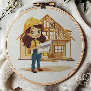 Op de afbeelding: Kruissteekborduurwerk in een houten hoepel met een cartoonafbeelding van een vrouwelijke bouwvakker die bouwtekeningen vasthoudt voor een huisframe. De werknemer draagt een gele helm en een blauwe overall. Het ontwerp staat tegen een witte achtergrond.