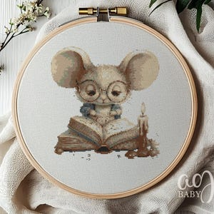 May include: Cross-stitch embroidery of a mouse wearing glasses, reading a book, and sitting in front of a lit candle. The artwork is framed in a wooden hoop. The mouse is light brown with large ears and is wearing round glasses.