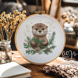 May include: A cross-stitch embroidery hoop with a white background featuring a brown and white otter holding a sprig of green leaves. The otter is surrounded by green leaves and pine cones. The hoop is on a wooden table with a book and a blanket.
