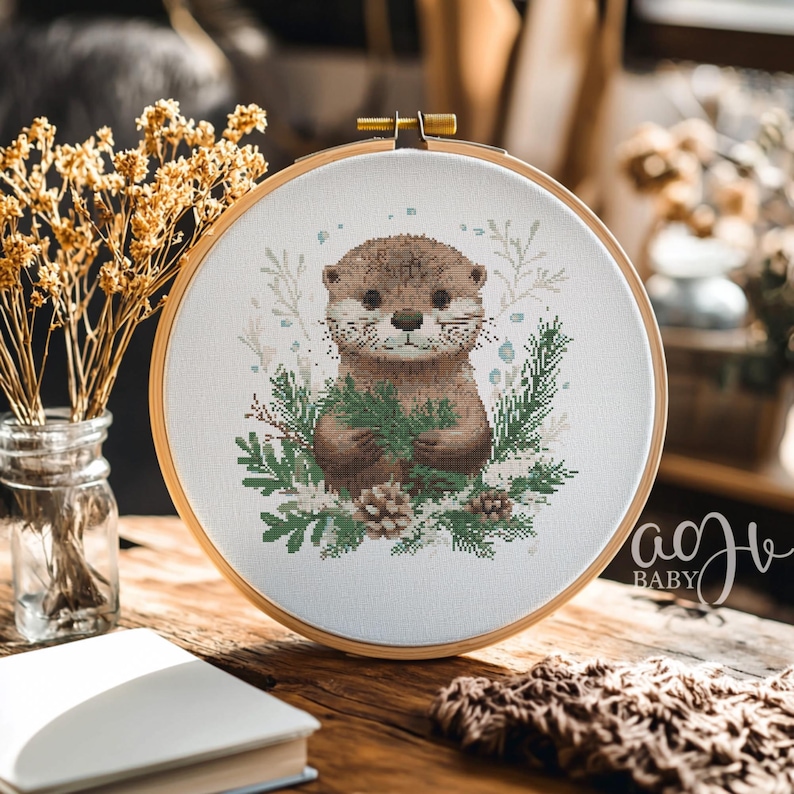 May include: A cross-stitch embroidery hoop with a white background featuring a brown and white otter holding a sprig of green leaves. The otter is surrounded by green leaves and pine cones. The hoop is on a wooden table with a book and a blanket.