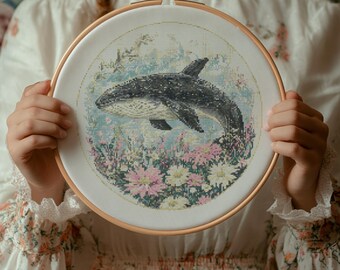 Può includere: Un cerchio da ricamo bianco con un disegno a punto croce di un'orca in bianco e nero che nuota in un campo di fiori rosa e bianchi.