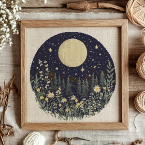 May include: A cross-stitch pattern featuring a full moon, stars, and a forest scene with flowers. The design is circular and framed in a dark wood frame.