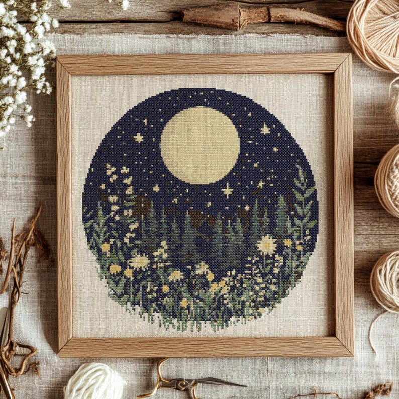 May include: A cross-stitch pattern featuring a full moon, stars, and a forest scene with flowers. The design is circular and framed in a dark wood frame.