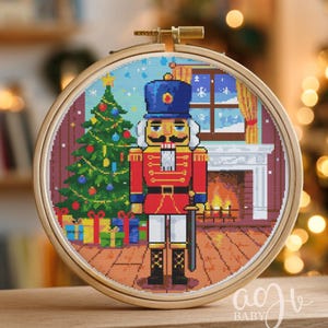 May include: A cross-stitch embroidery hoop featuring a pixelated design of a Nutcracker soldier in a room with a Christmas tree, fireplace, and window. The Nutcracker wears a red uniform and a blue hat. The hoop is made of wood.